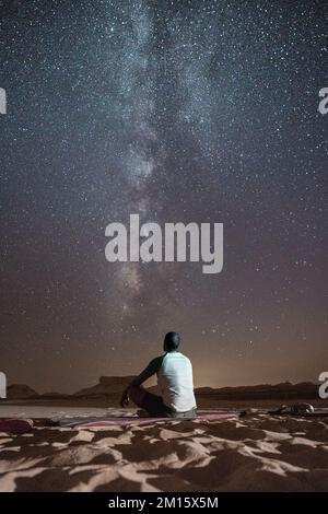 Vista posteriore di anonimo maschio nero seduto su un terreno sabbioso in Egitto e ammirando il cielo stellato spettacolare di notte Foto Stock