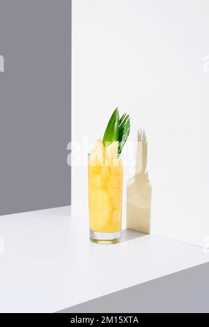 Bicchiere pieno di succo d'ananas freddo con foglie verdi e cannuccia posto su un tavolo illuminato da sole su sfondo bianco Foto Stock