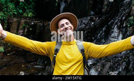 Felice maschio turista in cappello e maglione giallo con zaino mentre ammirando il paesaggio pittoresco Foto Stock