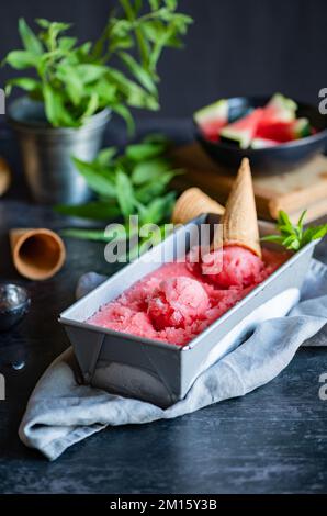 Da sopra cornetto posto in contenitore metallico di delizioso sorbetto di anguria su tovagliolo grigio e tavolo il giorno d'estate Foto Stock