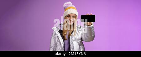 Ragazza bionda sicura e accogliente con giacca scintillante all'aperto in argento smartphone hat hold orizzontale che mostra il display del telefono cellulare assertivo Foto Stock