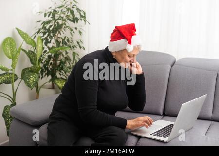 Donna anziana sorridente che indossa il cappello di Santa a Natale a digitare sul notebook da casa in contatto con la famiglia o gli amici. Foto Stock
