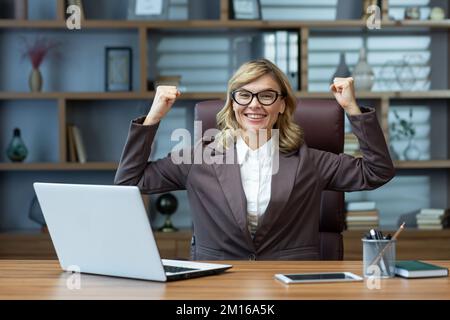 Donna d'affari matura di successo che tiene le mani in su gesto di fiducia e di forza sorridendo e guardando la fotocamera, capo femmina che lavora all'interno di un ufficio moderno indossando occhiali utilizzando un computer portatile. Foto Stock