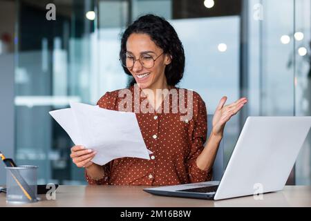 Giovane donna latino-americana sorridente scrittrice, giornalista siede in ufficio al tavolo con un computer portatile, tiene i documenti in mano, scrive un articolo, un libro. Foto Stock