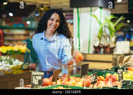 Stile di vita sano. Giovane bella studentessa latino-americana, vegetariana sceglie e compra le mele nel supermercato, mette la frutta in una borsa ecologica. Guarda la telecamera, sorride. Foto Stock