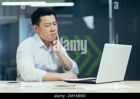 Mal di denti al lavoro. Un giovane asiatico siede in ufficio al tavolo, lavora su un computer portatile, tiene la guancia, sente un mal di denti. Foto Stock