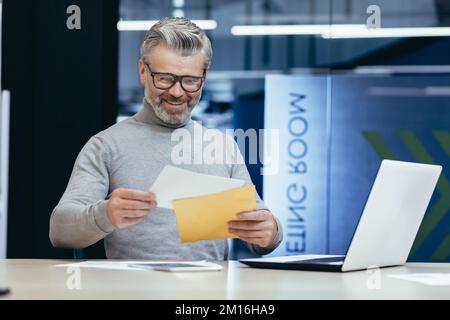 Maturo riuscito grigio haired businessman sorridente e felice ricevuto la notifica della lettera, capo maschio senior che lavora all'interno dell'ufficio usando il laptop che legge le buone notizie nella lettera. Foto Stock