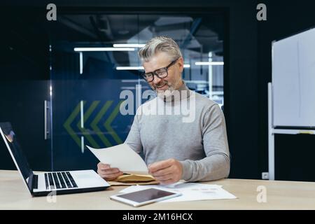 Maturo riuscito grigio haired businessman sorridente e felice ricevuto la notifica della lettera, capo maschio senior che lavora all'interno dell'ufficio usando il laptop che legge le buone notizie nella lettera. Foto Stock