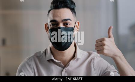 Etnia maschio studente uomo d'affari ispanico ragazzo in maschera medica proteggere da coronavirus focolaio in posa al chiuso mostrando pollici in su come gesto raccomandare Foto Stock