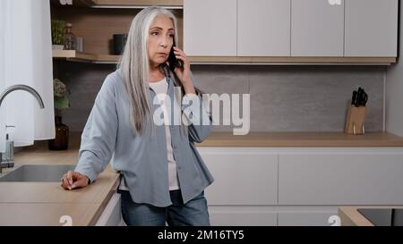 Sconvolto caucasico vecchia donna delusa anziana signora grigio-capelli nonna businesswoman stand in cucina parlare telefono cellulare risposta spiacevole chiamata rec Foto Stock