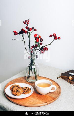 Tazza di tè e biscotti al cioccolato su server in legno con rami di rosa in vaso di vetro. Accogliente colazione invernale. Ancora vita. Foto Stock