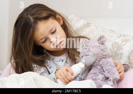 Preoccupata, senza sole, stanca bambina bruna abbracciando paziente giocattolo orso, guarendo con spray naso, sedersi a letto in camera da letto. Isolamento Lockdown, loneline Foto Stock