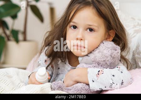 Triste depressa preoccupata, ragazza piccola brunetta senza sole che abbraccia il giocattolo dell'orso, healing con lo spruzzo del naso, sedersi sul letto e guardare la macchina fotografica. Malattia di capretto, virus AN Foto Stock