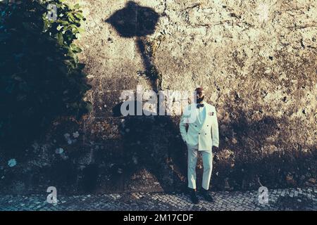 Alto uomo africano calvo e bearded vestito in un vestito bianco con un arco punteggiato di polka, occhiali e scarpe coordinate. Dietro di lui un muro di pietra marrone con un s Foto Stock