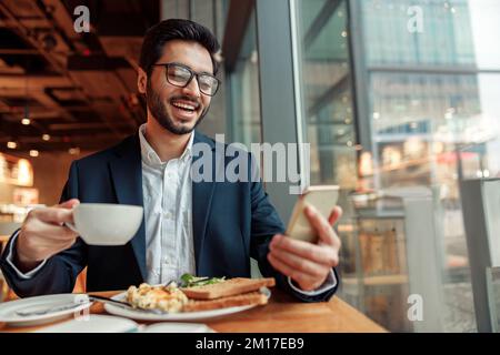Un uomo d'affari indiano sorridente che indossa un costume usa il telefono durante il pranzo nel bar. Sfondo sfocato Foto Stock