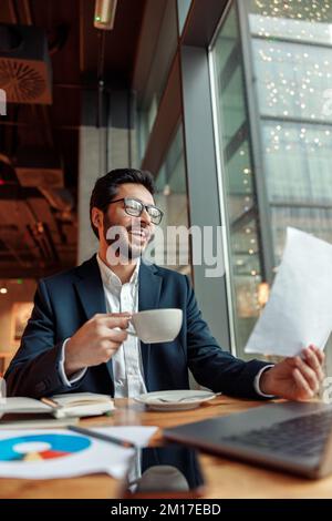 Uomo d'affari indiano sorridente che beve caffè e lavora con i documenti nel caffè Foto Stock