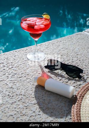 Rinfrescante cocktail rosso in piscina con crema solare, occhiali da sole e cappello di paglia Foto Stock