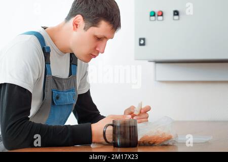 Pausa pranzo. Mangiare sul posto di lavoro durante il lavoro. L'uomo caucasico in tuta da lavoro siede al tavolo nella sala di produzione e mangia dal contenitore. Il lavoratore ha pranzo in sala pausa. Foto Stock