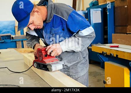 Falegname in abiti da lavoro lavora legno con macinacaffè. Lucidatura della superficie del legno. Uomo di mezza età lavora al banco di lavoro in Falegnameria officina. Flusso di lavoro reale. Foto Stock