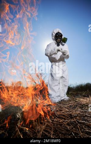 Persona irriconoscibile che brucia erba secca vecchia in campo. Ecologista che tiene matchbox e di messa fuoco per asciugare erba sotto il cielo blu. Ricercatore scienziato che indossa tuta protettiva contro le radiazioni e maschera a gas. Foto Stock