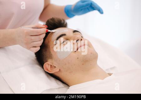 Estetista applicare la maschera di argilla sul viso dell'uomo nel salone termale. Primo piano del lato maschio durante la procedura. Pimples e blackheads sulla pelle. Acne Foto Stock