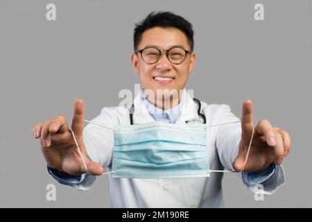 Buon terapista asiatico di mezza età in bianco cappotto e occhiali mette maschera sulla macchina fotografica Foto Stock