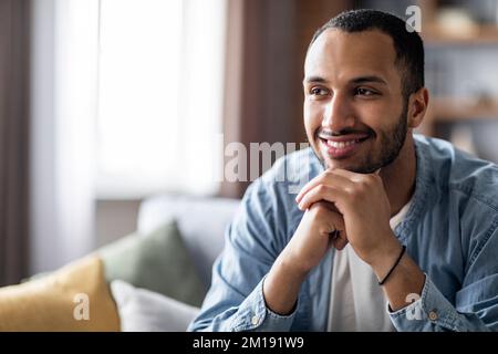 Un bel ragazzo nero sorridente seduto sul divano a casa e guardando lontano Foto Stock