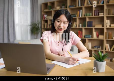 Studentessa asiatica positiva che studia davanti al portatile e prende appunti, indossando cuffie wireless, spazio copia Foto Stock