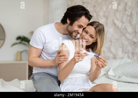 Ritratto di felice coppia caucasica che tiene test di gravidanza positivo, seduto a letto e godendo di buone notizie Foto Stock