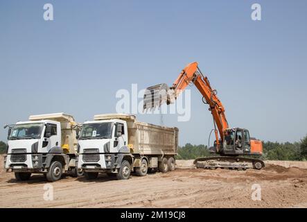 escavatore cingolato arancione e due dumper da cantiere grigi nel processo di carico e trasporto del terreno in una cava contro un cielo blu. Commerc Foto Stock