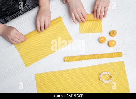 Due capretti fanno le candele rotolate naturali della cera d'api dal foglio a nido d'ape pressato della cera d'api nel paese all'interno. Concetto di hobby. Due candele accese e accese. Foto Stock
