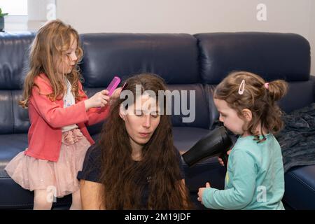 Una madre e le sue due figlie giocano e interagiscono insieme, con una ragazza che pettina i capelli bagnati e l'altra che asciuga i capelli con un asciugacapelli Foto Stock