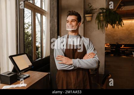 Giovane cameriere bianco che indossa grembiule e maschera facciale in piedi davanti al computer mentre lavora in un bar Foto Stock