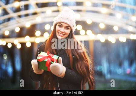 Giovane donna in un cappello a maglia, che sorride e tiene un regalo di Natale nelle sue mani sullo sfondo di neve e luci di Natale Foto Stock