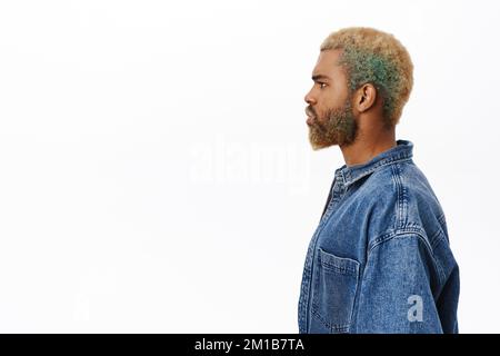 Profilo ritratto di elegante giovane afro-americano, ha barba gialla e acconciatura, indossa giacca denim, guarda a sinistra, si erge su sfondo bianco Foto Stock