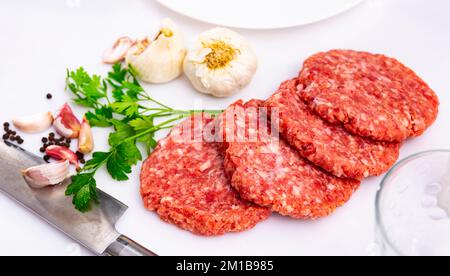 Hamburger crudi di maiale se si tratta di carne macinata su un piatto decorato con pepe, aglio e prezzemolo su sfondo bianco Foto Stock