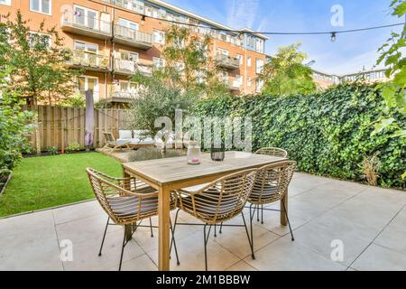 un patio con sedie, tavolo e ombrelloni a terra di fronte ad un edificio di appartamenti circondato da alberi Foto Stock