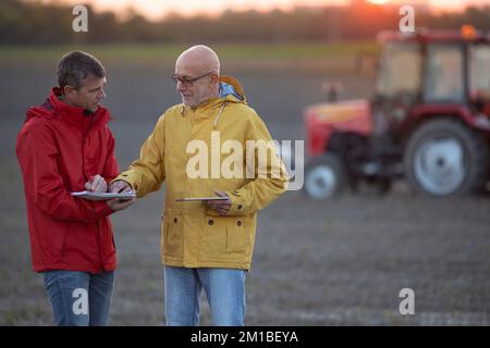 Due agricoltori anziani scuotono le mani sul campo al tramonto in autunno, mentre il trattore lavora in background Foto Stock
