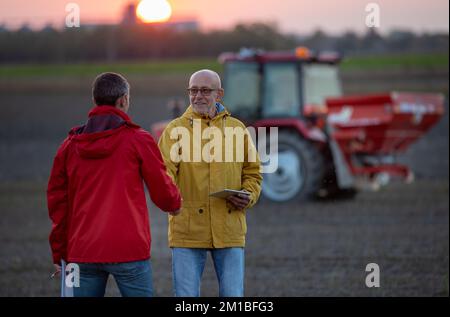 Due agricoltori anziani scuotono le mani sul campo al tramonto in autunno, mentre il trattore lavora in background Foto Stock
