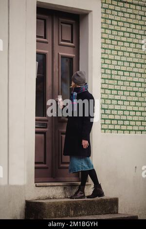 Una donna in un cappotto sul portico di una casa bussa alla porta. Battito in testa. Foto Stock