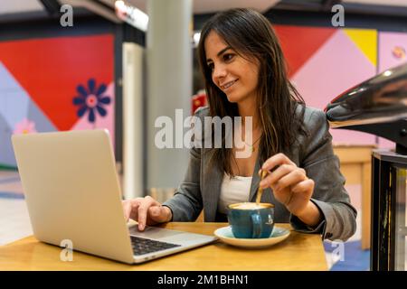 Imprenditrice femminile positiva che mescola latte in tazza e dati di navigazione su netbook mentre si siede al tavolo del caffè e lavora in remoto in centro commerciale Foto Stock