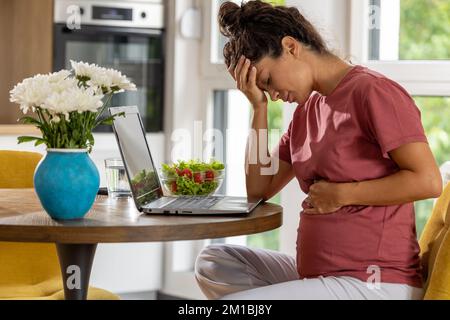 Donna incinta seduta al tavolo di fronte al computer portatile e sensazione di nausea Foto Stock