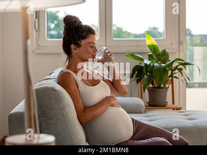 Bella donna incinta seduta sul divano, riposo e acqua potabile sul divano in soggiorno Foto Stock