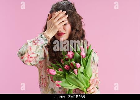 stressato donna moderna con lunghi capelli di brunette ondulate con bouquet tulipani isolato su rosa. Foto Stock