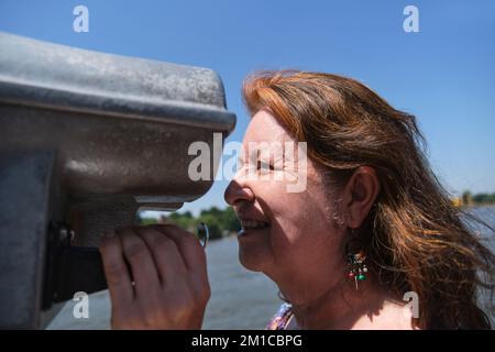 Ritratto di una donna matura ispanica che si diverte a guardare attraverso binocoli pubblici. Concetti di curiosità, osservazione, esplorazione, ricerca, viaggio. Foto Stock