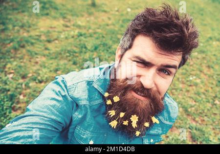 Ragazzo con minori fiori di celandina in barba scattando foto selfie. Hippster con allegro grimace sul viso scattando foto selfie. L'uomo ama la primavera, il verde Foto Stock