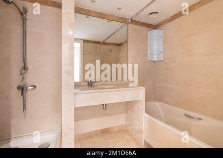 Bagno luminoso con pareti piastrellate e pavimenti in calde tonalità beige chiaro. Lavabo con grande specchio, vasca in acrilico bianco e zona doccia per prendere Foto Stock