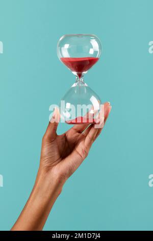 Crop ragazza afro-americana irriconoscibile che mostra clessidra con sabbia rossa su sfondo turchese Foto Stock