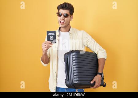 Giovane uomo ispanico che tiene valigia andare in vacanza estiva con passaporto italiano in shock faccia, guardando scettico e sarcastico, sorpreso con o Foto Stock