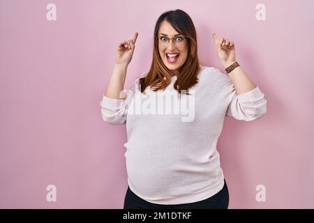 Donna incinta in piedi su sfondo rosa sorridente stupito e sorpreso e puntando in su con le dita e le braccia sollevate. Foto Stock
